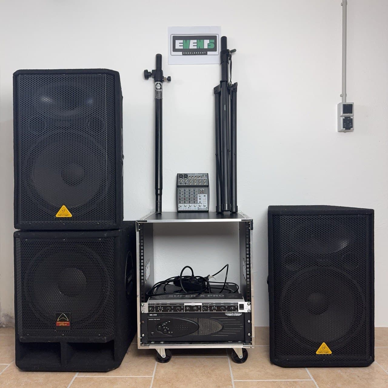 Audiogeräte-Setup mit zwei großen Lautsprechern, einem Mixer, Ständern und Kabeln, ordentlich organisiert auf einem Regal vor einer weißen Wand.