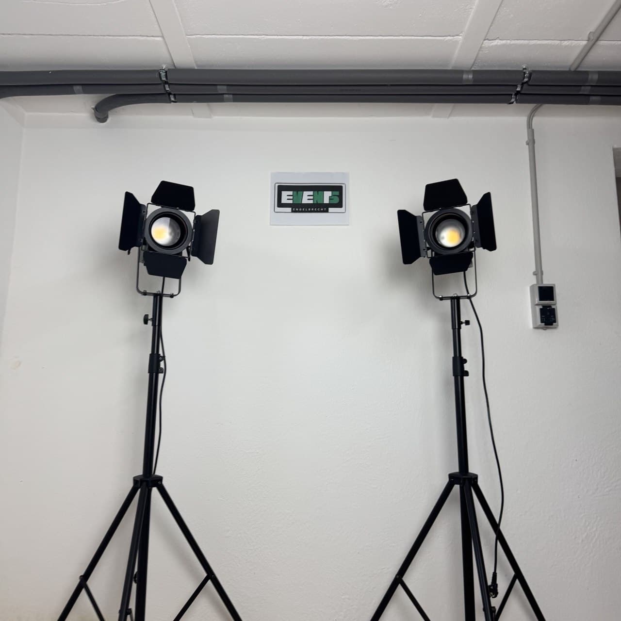 Zwei Studioleuchten auf Stativen beleuchten eine weiße Wand, über der sich ein Bedienfeld befindet. Kabelkanäle verlaufen entlang der Decke.