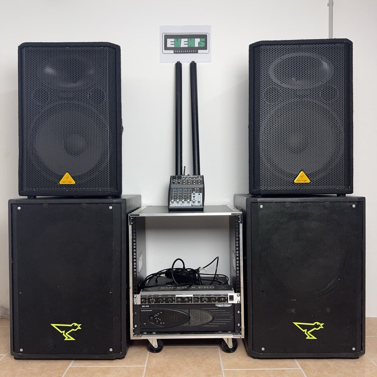 Zwei große schwarze Lautsprecher auf beiden Seiten einer Audiomischer- und Verstärkeranlage mit einem Logo-Schild an der Wand darüber.