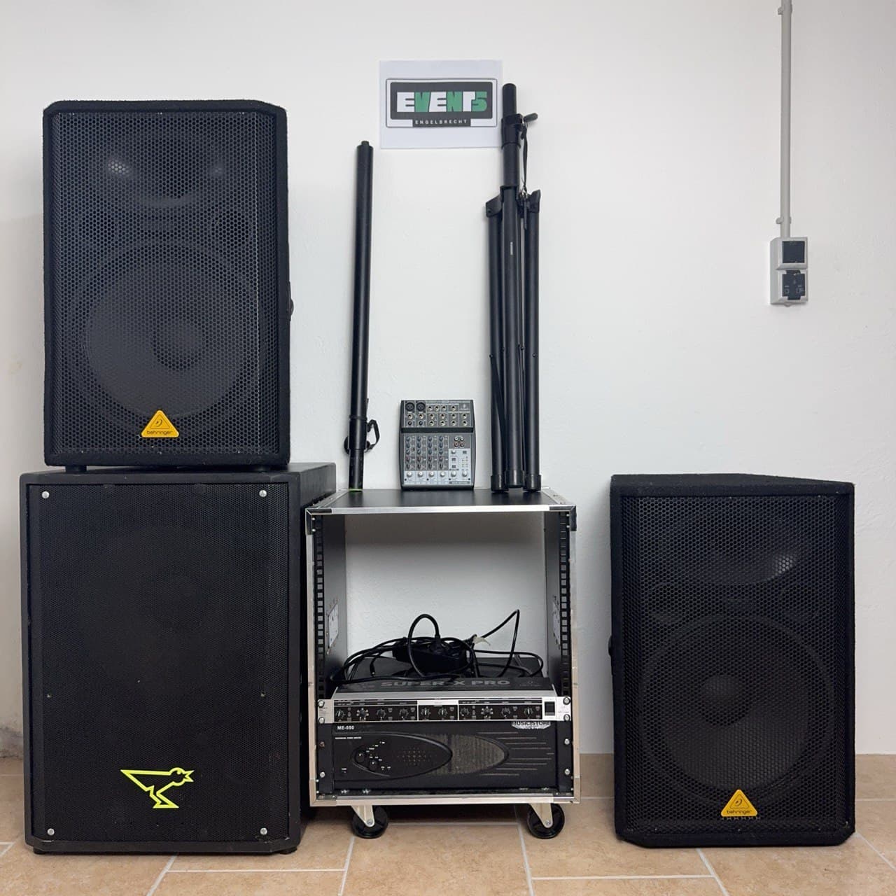 Aufbau einer Audioausrüstung mit großen Lautsprechern, einem Audiomischer und Kabeln auf einem Fliesenboden. An der Wand darüber befindet sich ein Schild.