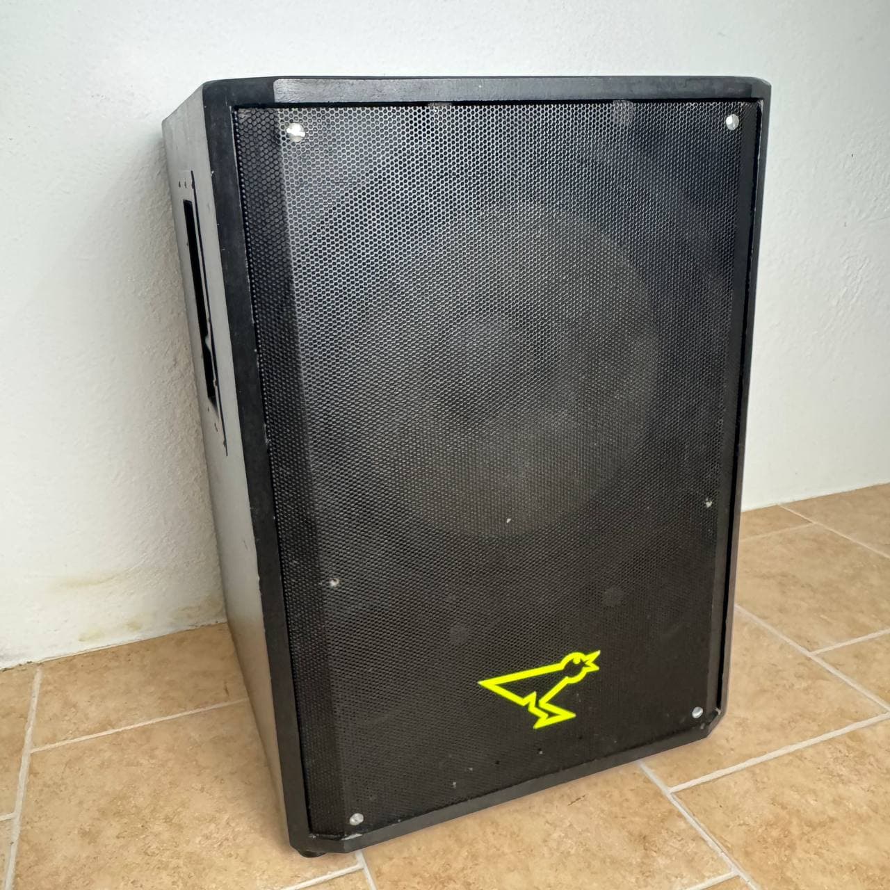 Schwarzer Lautsprecher mit Netzfront und neongrünem Logo, platziert auf einem hellbraunen Fliesenboden vor einer hellen Wand.