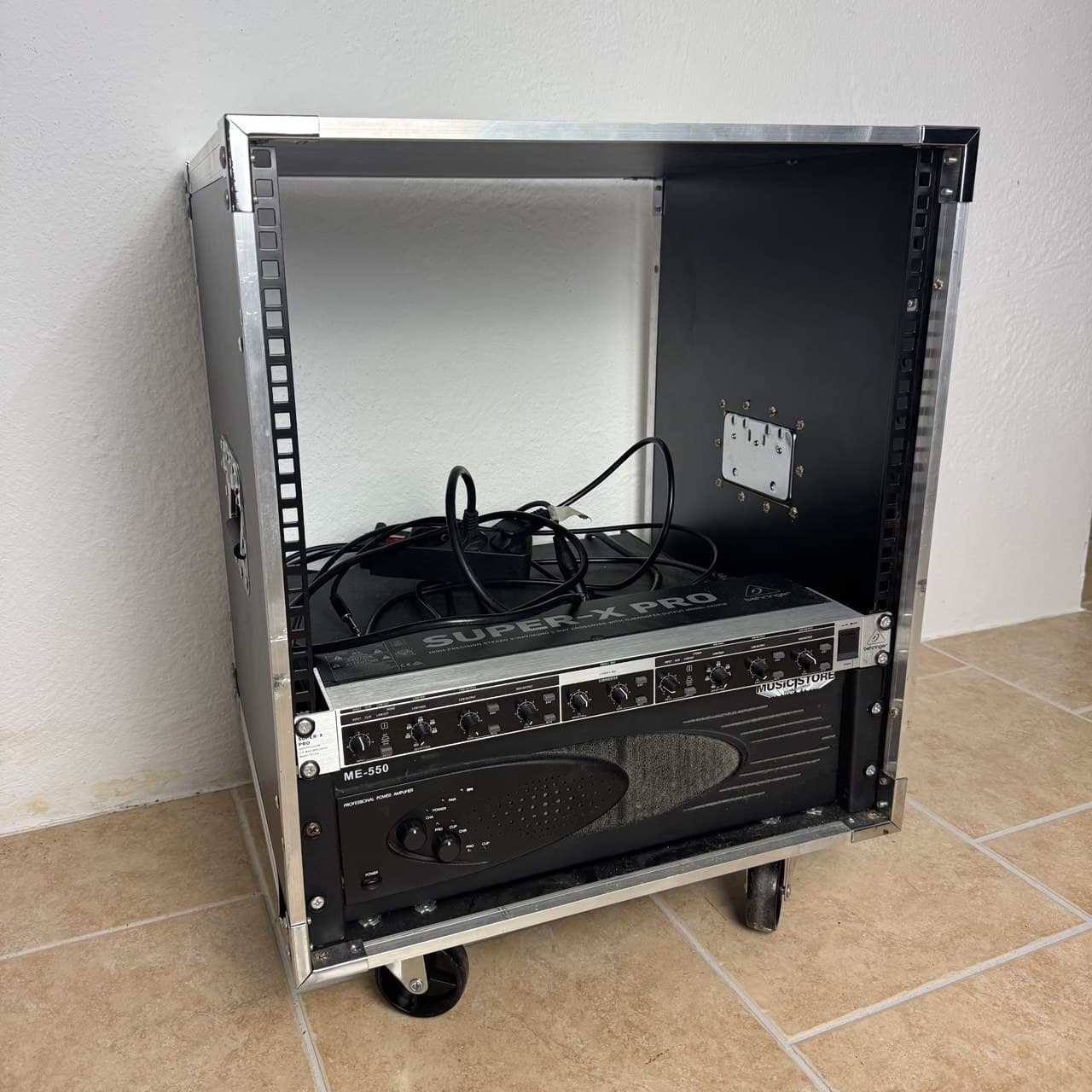Ein Metall-Rack-Gehäuse mit Audiogeräten, darunter eine Behringer Super-X Pro-Frequenzweiche und ein ME-200-Verstärker, auf einem Fliesenboden.
