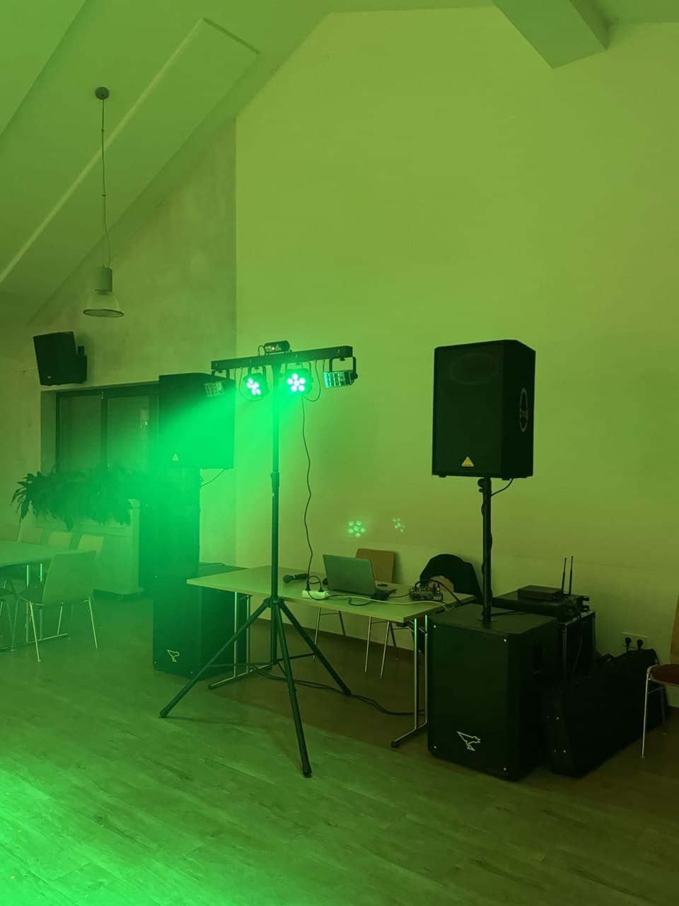 Raum mit grünen Bühnenlichtern, Lautsprechern und DJ-Equipment auf Tischen an der Wand. Im Hintergrund stehen leere Stühle und Pflanzen.