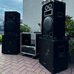 Audioanlage im Freien mit großen schwarzen Lautsprechern und einem Tonmischer auf einer gemauerten Terrasse, umgeben von viel Grün und einer Gebäudemauer.