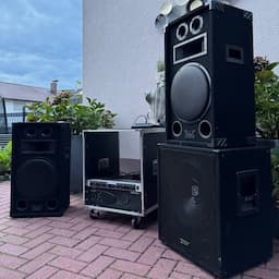 Audioanlage im Freien mit großen schwarzen Lautsprechern und einem Tonmischer auf einer gemauerten Terrasse, umgeben von viel Grün und einer Gebäudemauer.