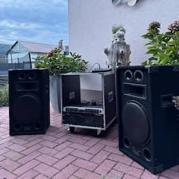 Audioanlage im Freien mit großen schwarzen Lautsprechern und einem Tonmischer auf einer gemauerten Terrasse, umgeben von viel Grün und einer Gebäudemauer.