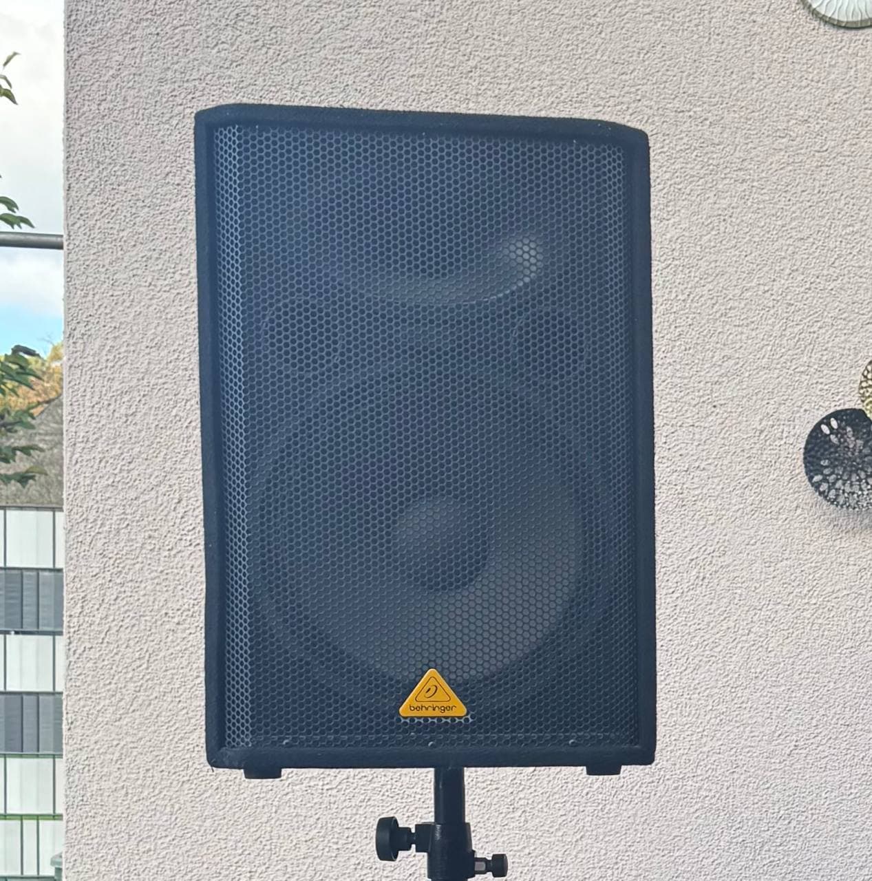 Schwarzer Behringer-Lautsprecher auf einem Ständer vor einer beigen Wand, mit kreisförmigen Verzierungen und teilweisem Blick auf ein Fenster.