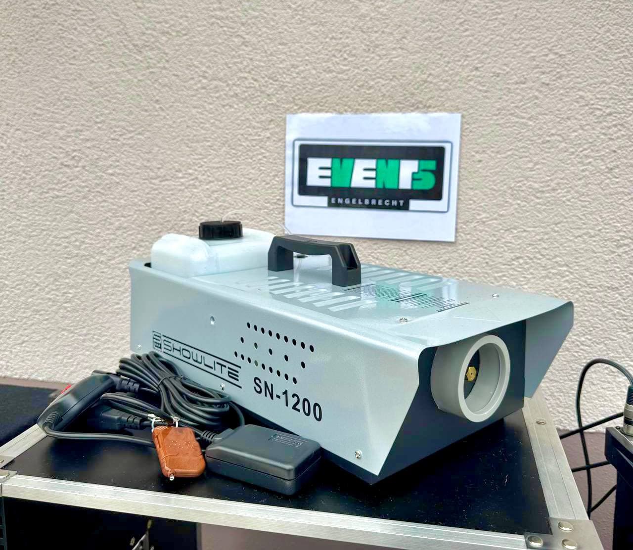 Eine Showlite SN-1200 Nebelmaschine auf einem Tisch, mit Kabeln und einer Fernbedienung. An der Wand dahinter hängt ein Schild mit der Aufschrift „EVENTS“.