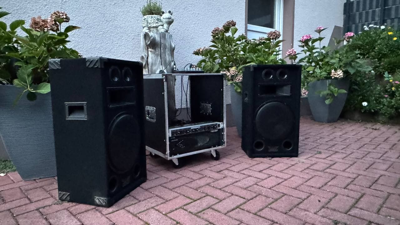 Außenanlage mit großen schwarzen Lautsprechern und Audiogeräten auf einer gemauerten Terrasse, umgeben von Topfpflanzen und einem weißen Gebäude.