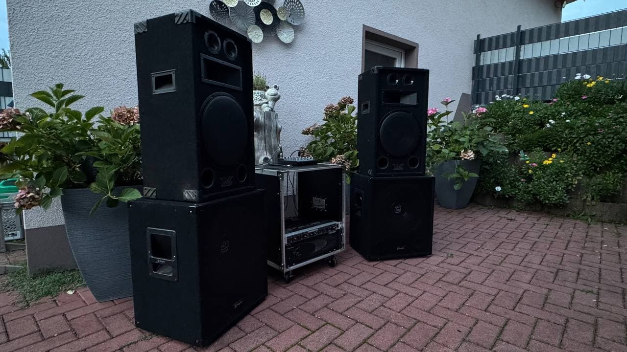 Außenanlage mit großen schwarzen Lautsprechern und Audiogeräten auf einer gemauerten Terrasse, umgeben von Topfpflanzen und einem weißen Gebäude.
