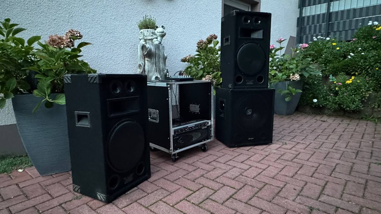 Außenanlage mit großen schwarzen Lautsprechern und Audiogeräten auf einer gemauerten Terrasse, umgeben von Topfpflanzen und einem weißen Gebäude.