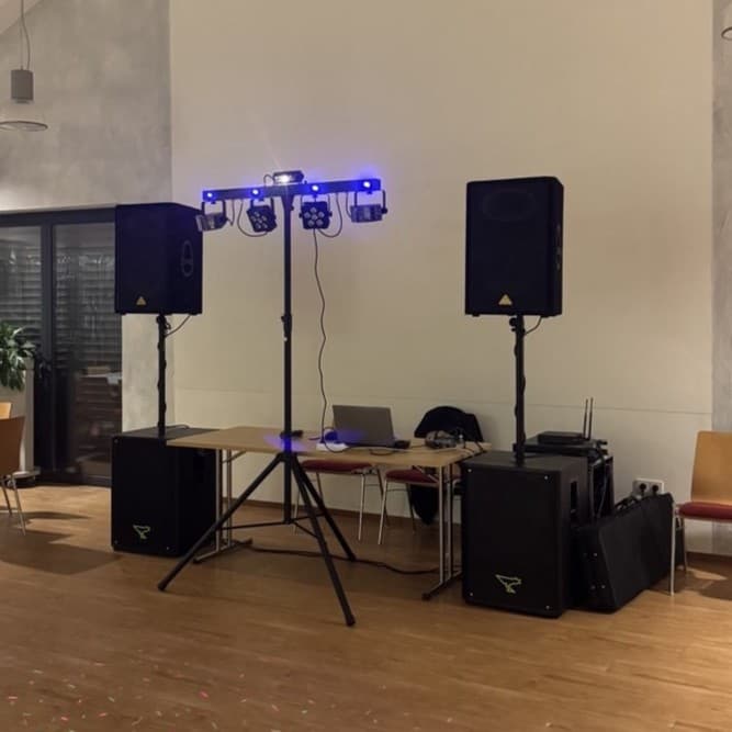 DJ-Setup mit Lautsprechern, Beleuchtung und einem Laptop auf einem Tisch in einem Raum mit Holzböden und Stühlen.